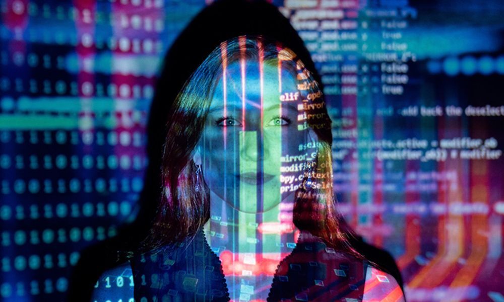 A woman in a hood is standing in front of a computer screen.