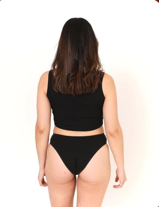 Mujer de pie de espaldas a la cámara con un top corto negro y ropa interior negra sobre un fondo blanco.