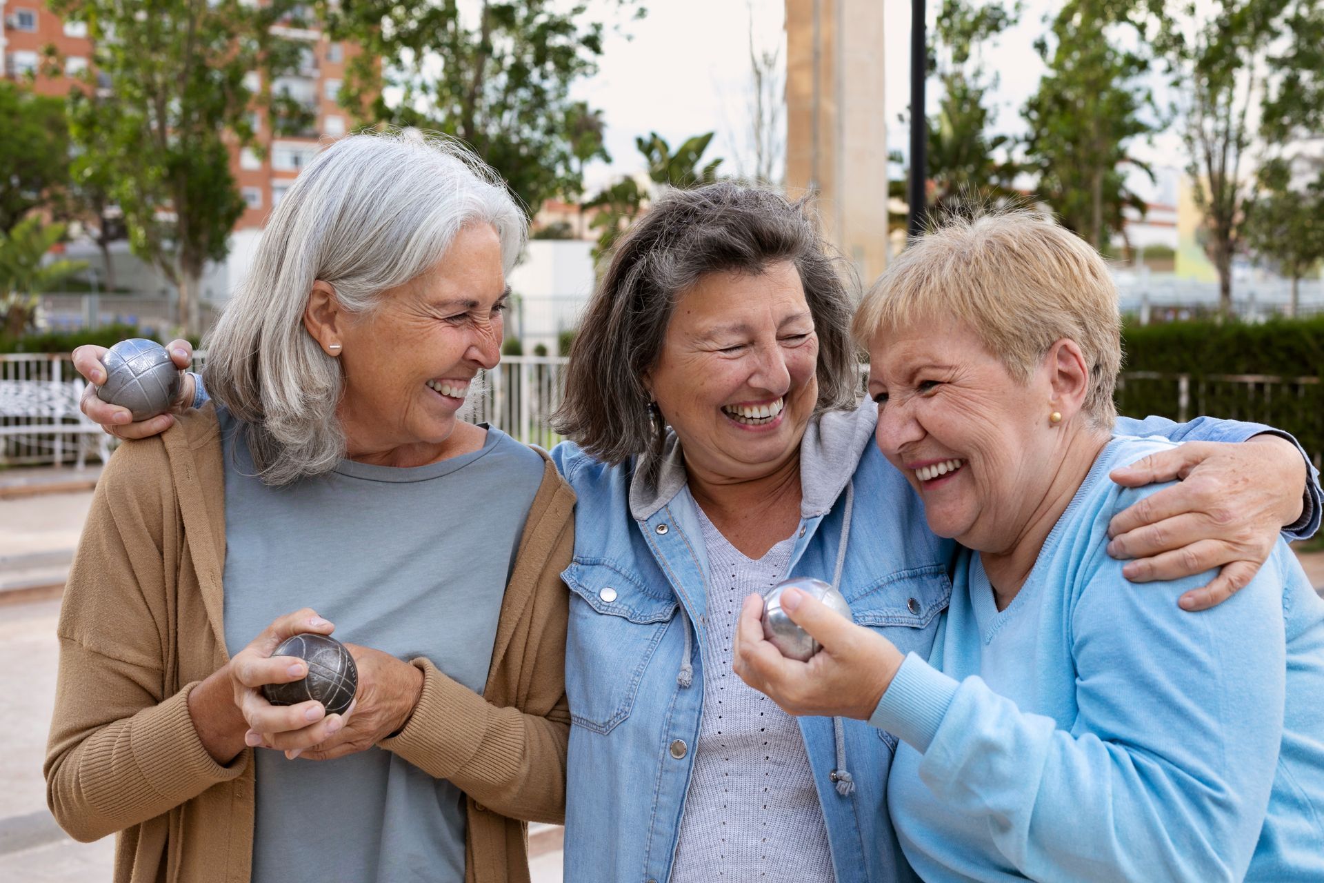 Three women laugh together, holding metal balls outdoors.