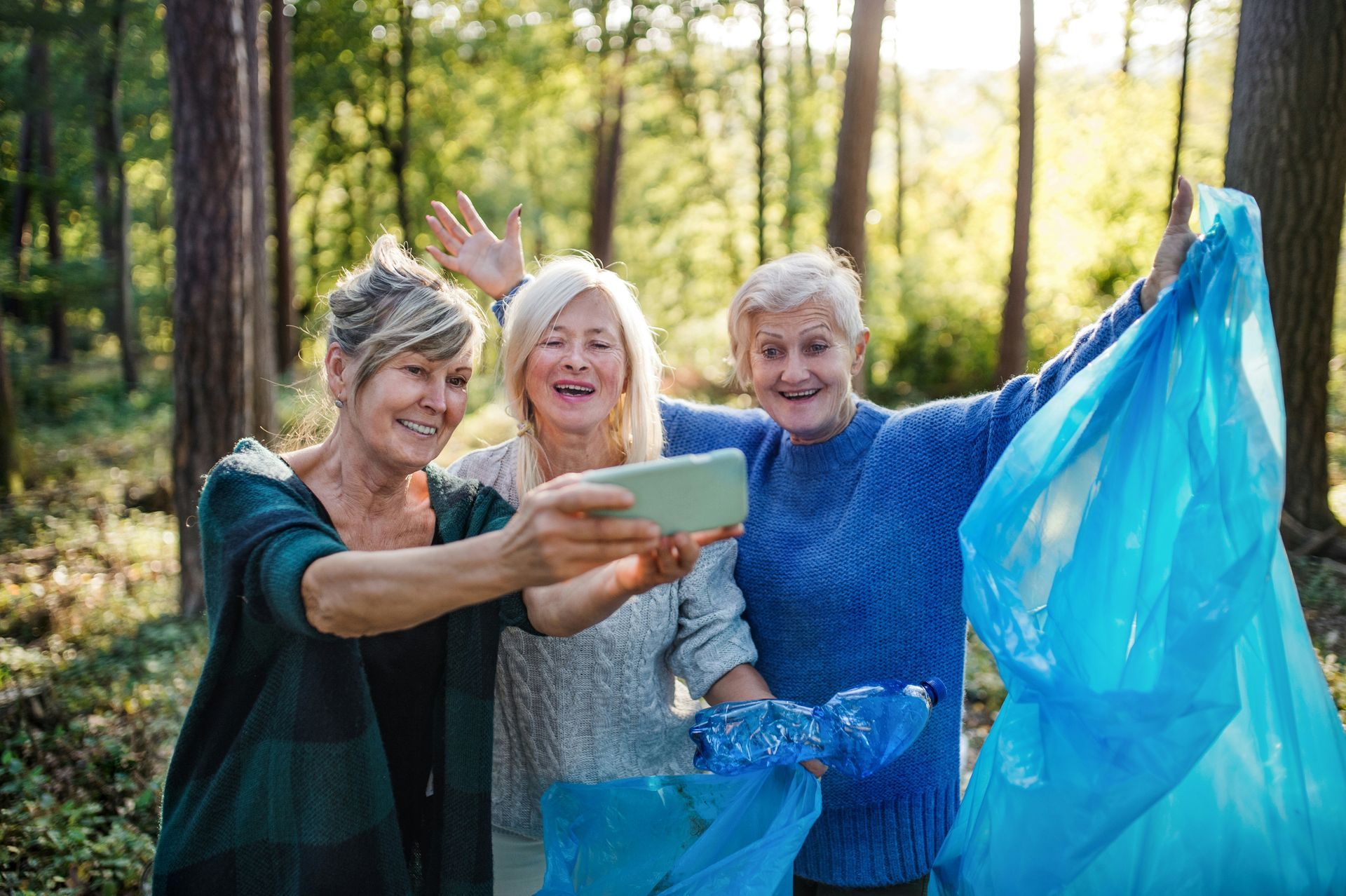 Three women smiling, taking a selfie while holding blue trash bags in a forest.