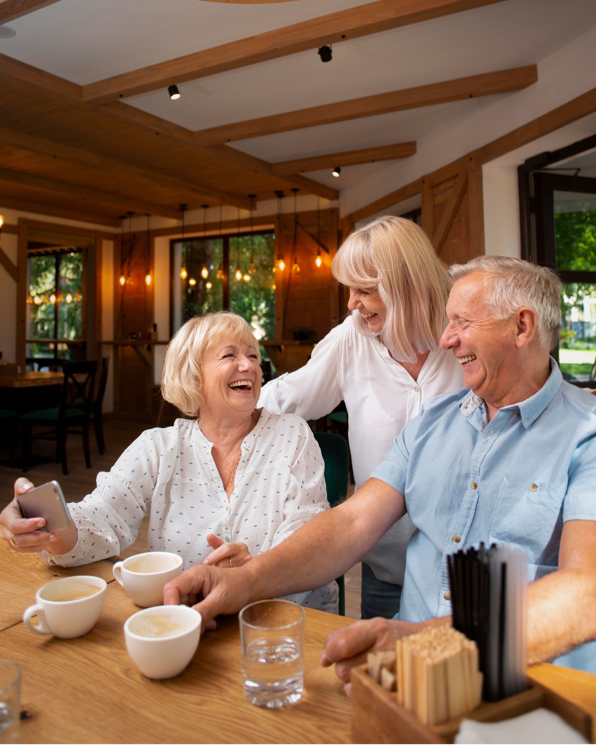 Three people laughing at a table. Woman takes selfie with phone. Coffee cups present. Wooden beams and interior.