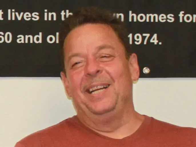 Man in a red shirt smiles and laughs, looking toward the camera. Black background with text.