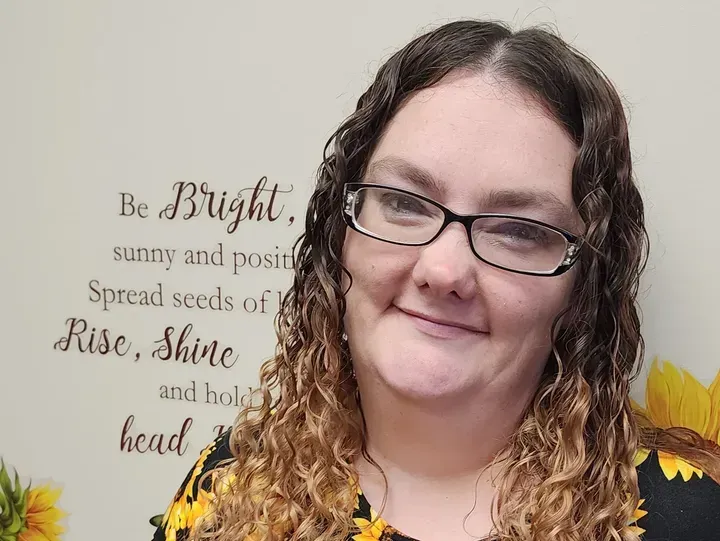 Woman with glasses smiling, against a wall with inspirational words and sunflower accents.