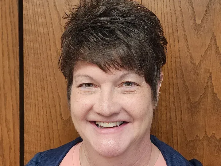 Woman with short, dark hair, smiling broadly in front of a wood-paneled wall.