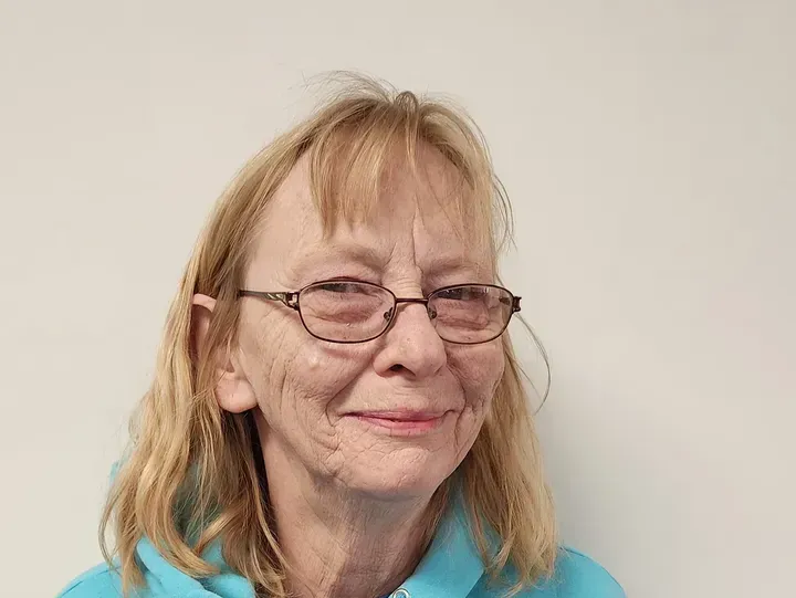 Woman with glasses, blond hair, smiling, wearing a blue shirt, in front of a white wall.