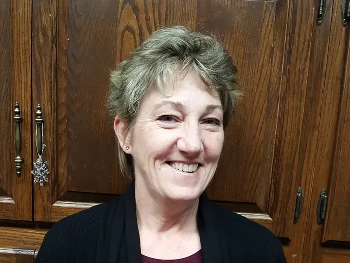 Woman smiling, short blonde hair, black sweater, in front of wooden cabinets.