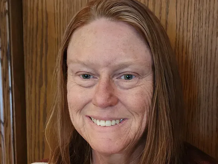 Woman with reddish-blonde hair smiling at the camera, wrinkles visible. Wooden door background.