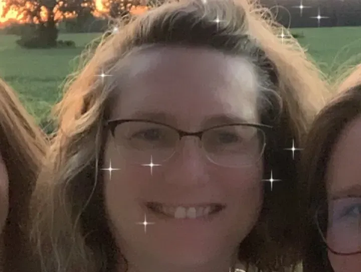 Woman with glasses smiles; outdoors, blurred background, sparkly effects.