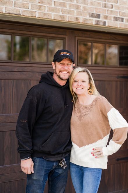 Two people standing together outdoors in front of a garage, smiling at the camera.