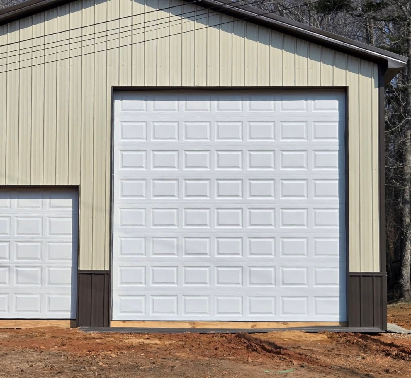 Two white garage doors on a beige metal building with dark trim and a dirt foreground