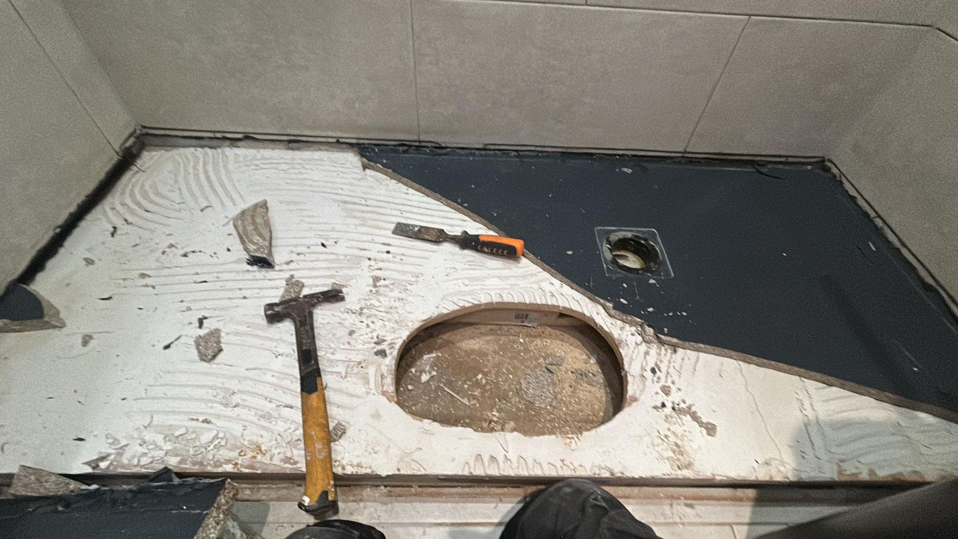 Bathroom floor under renovation with a toilet opening, hammer, tools, and debris.