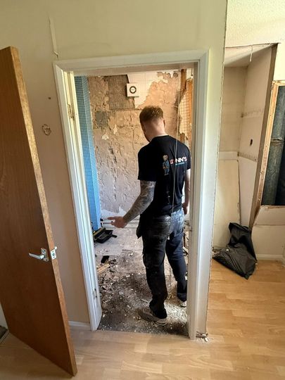 Worker standing in a doorway amid a room renovation, with debris on the floor and exposed wall behind.