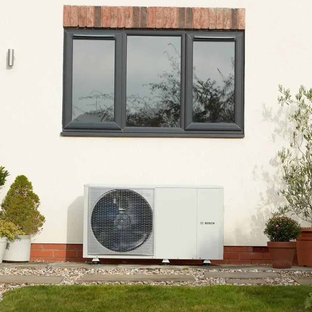 Outdoor wall-mounted heat pump beneath a three-pane window on a white house exterior