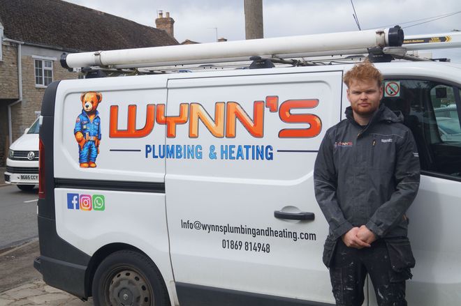 Man standing beside a white Wynn’s Plumbing & Heating van with company logo and phone number.