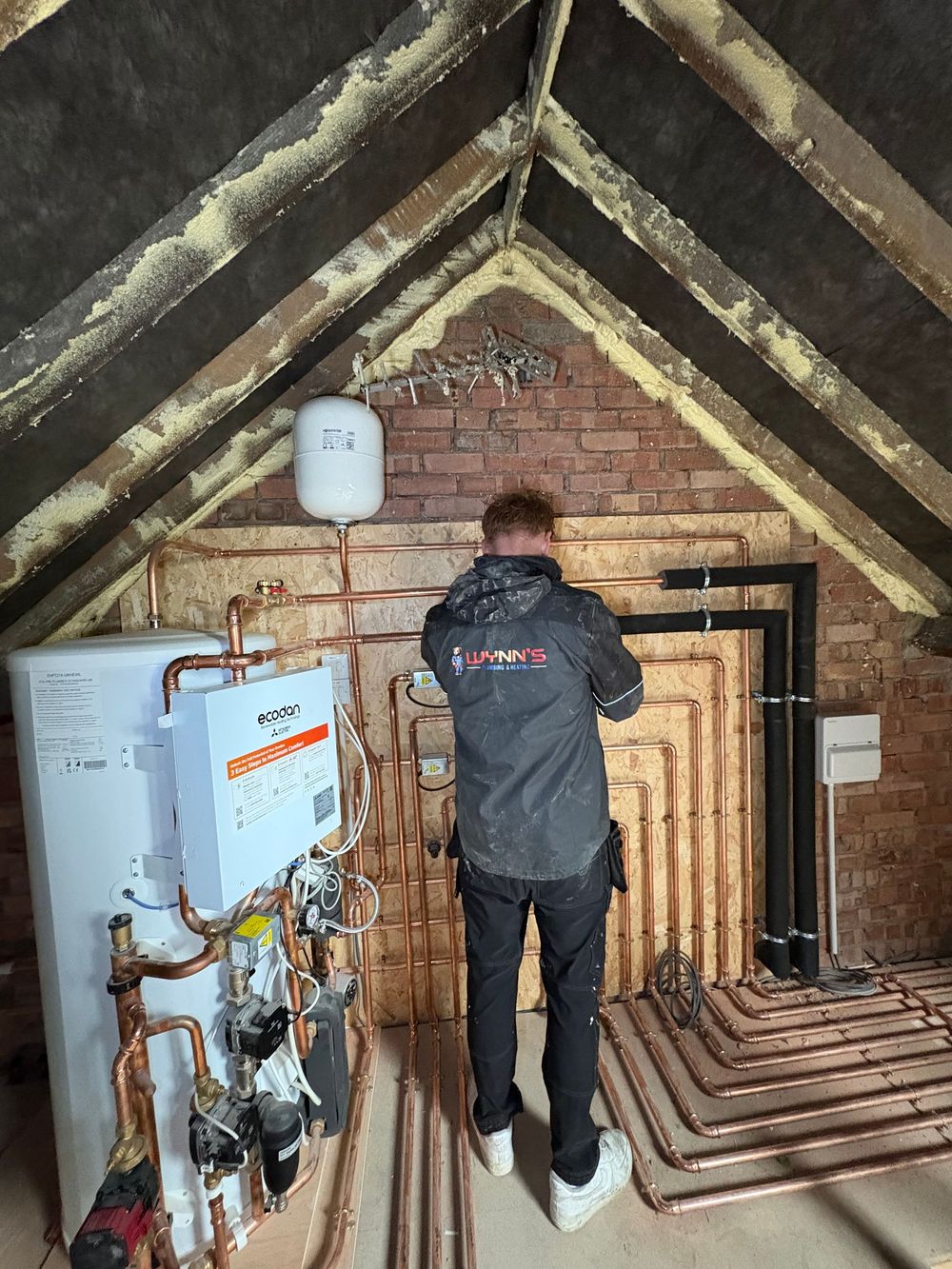 Worker inspecting exposed plumbing and heating pipes in an attic with brick walls and sloped beams