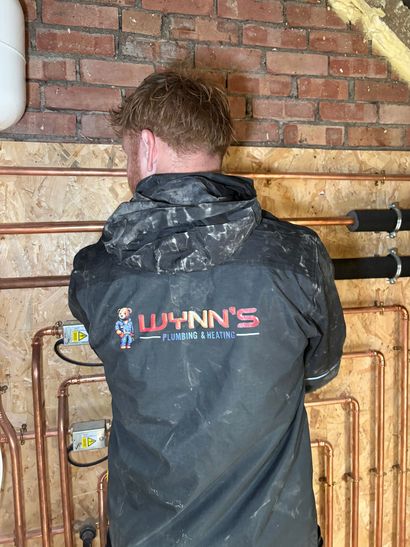 Technician in black Wynn’s jacket inspecting pipes against a brick wall in a boiler room