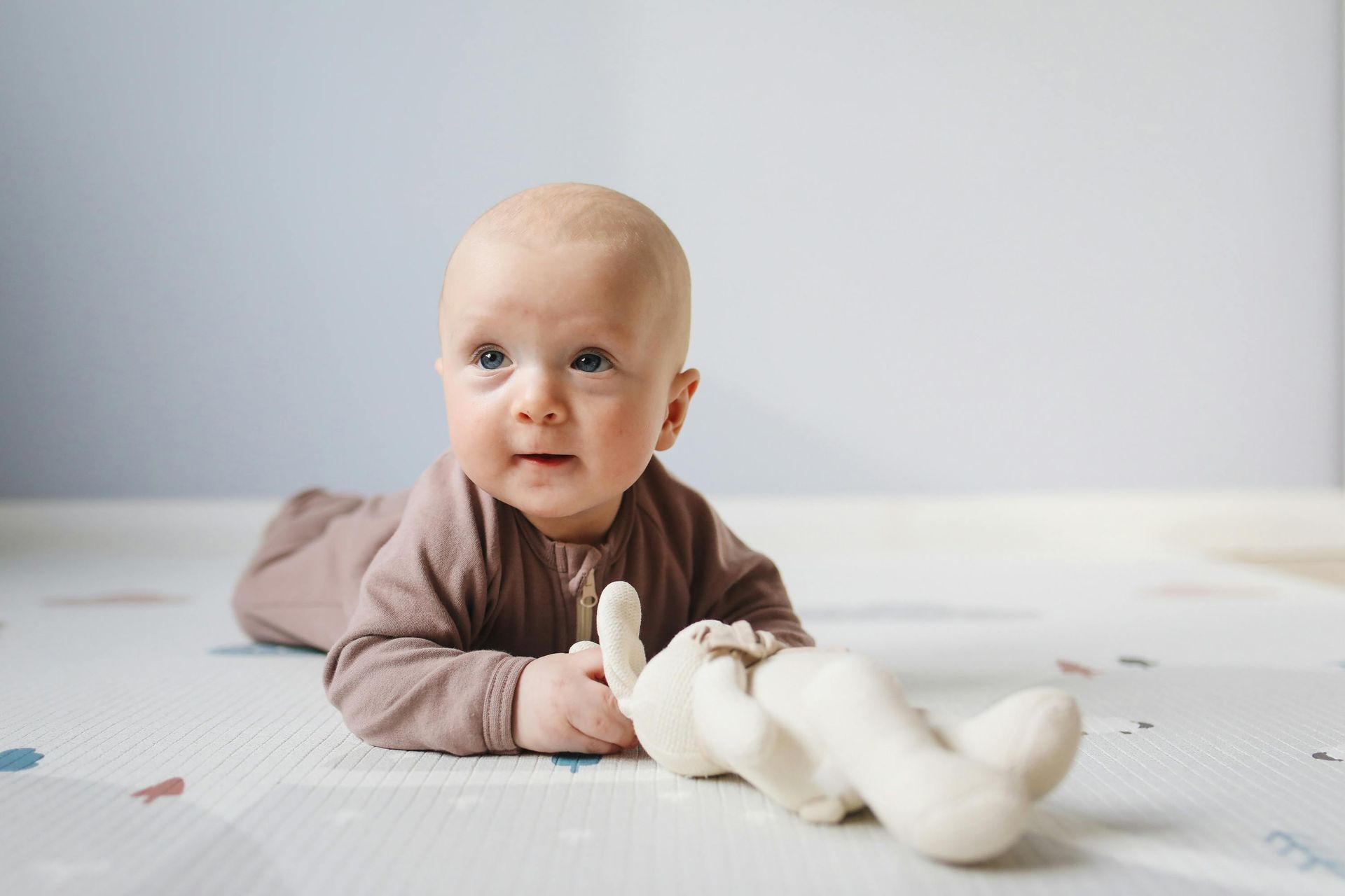 Baby on stomach, looking up, light brown outfit, toy bunny on playmat.