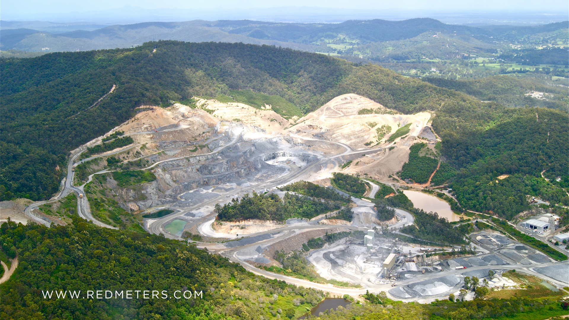 An aerial view of a large quarry carved into a mountainous area, surrounded by lush green trees.