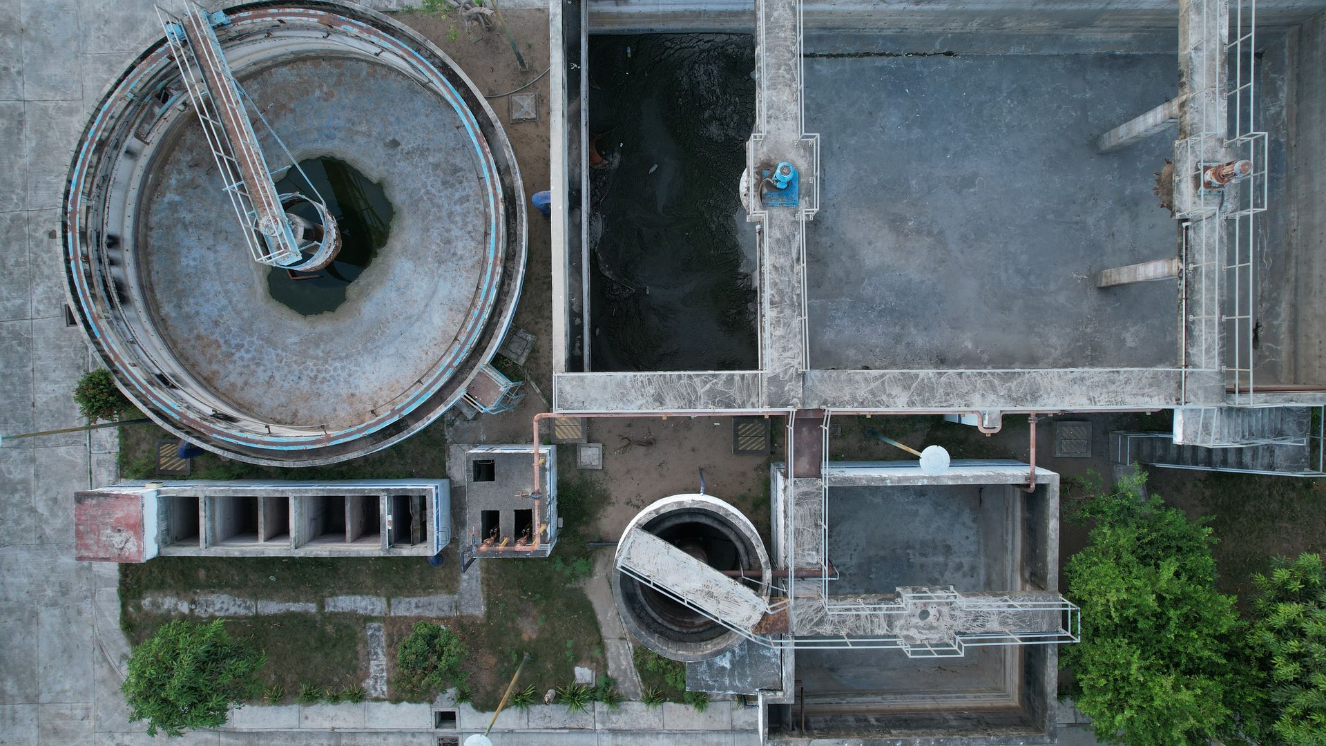 Aerial view of a wastewater treatment plant with circular tanks, rectangular basins, and concrete structures. Green foliage surrounds the plant.