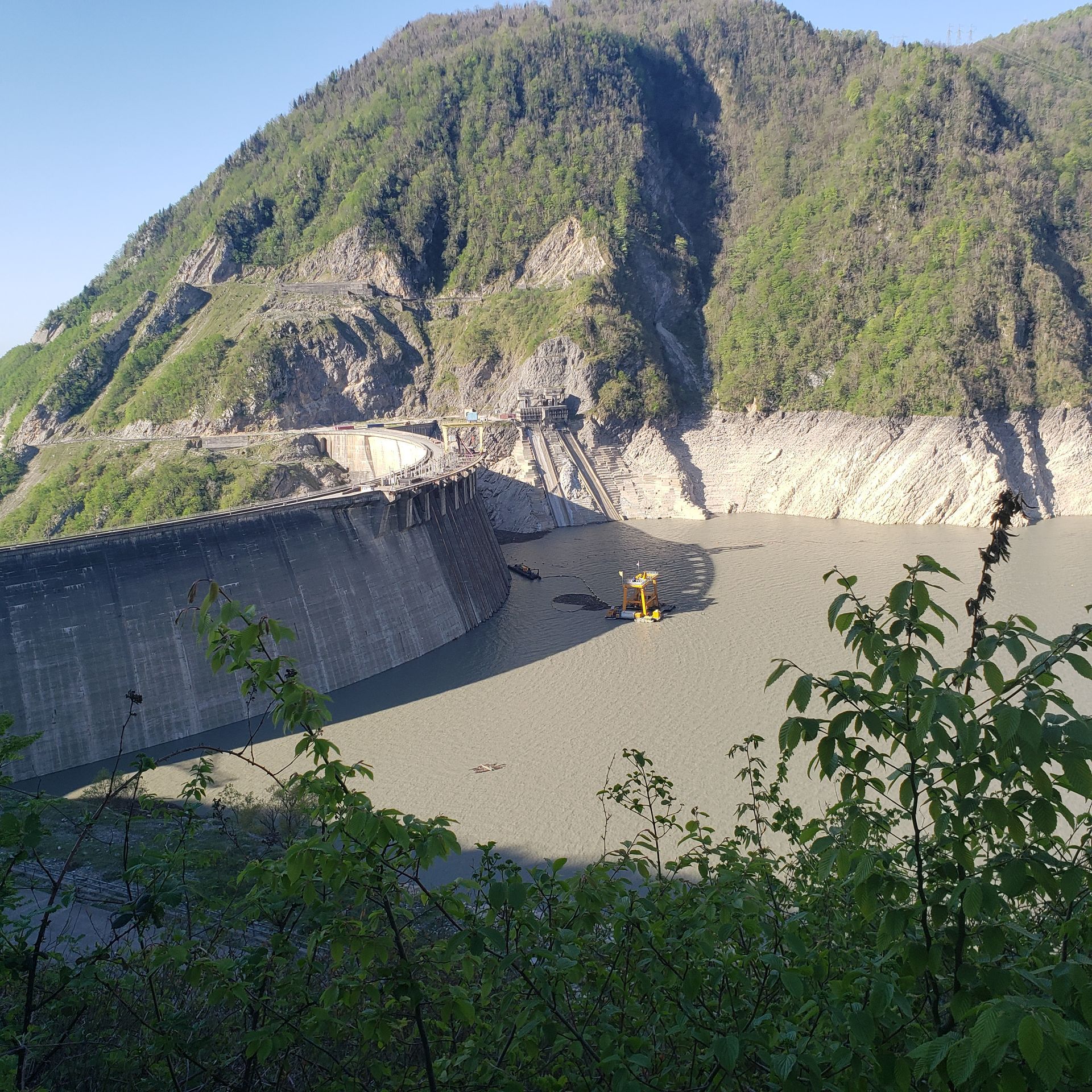 Curved dam spans a lake, backed by a large, green mountain. Some construction machinery is visible on the water.