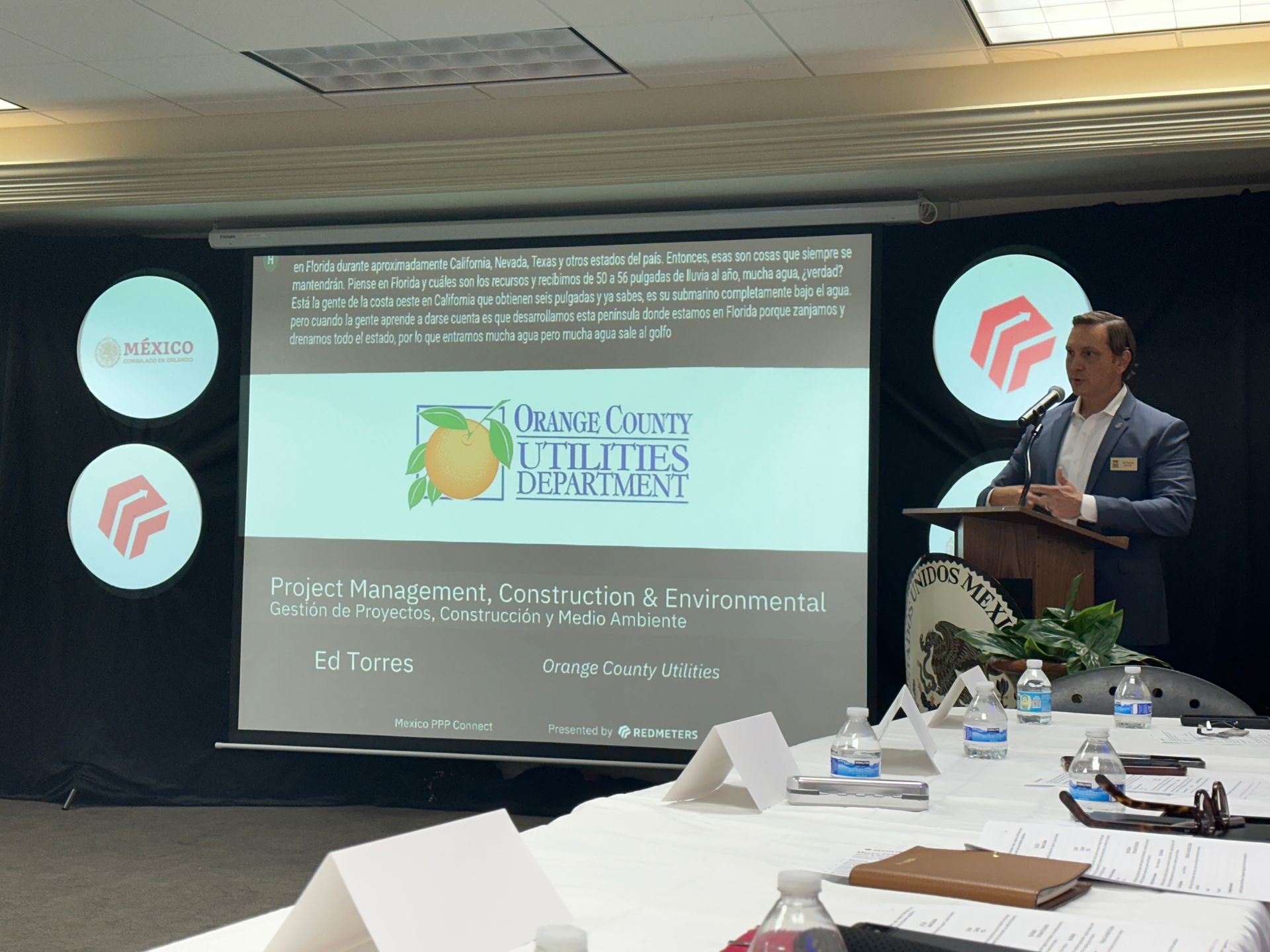 Man speaking at a podium in front of a presentation screen with the logo of the Hernando County Utilities Department. A table with documents and water bottles is in the foreground.