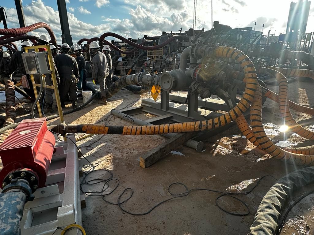 Workers connecting hoses to machinery, likely in an industrial setting. Yellow and black hoses are prominent, along with equipment and debris under a cloudy sky.