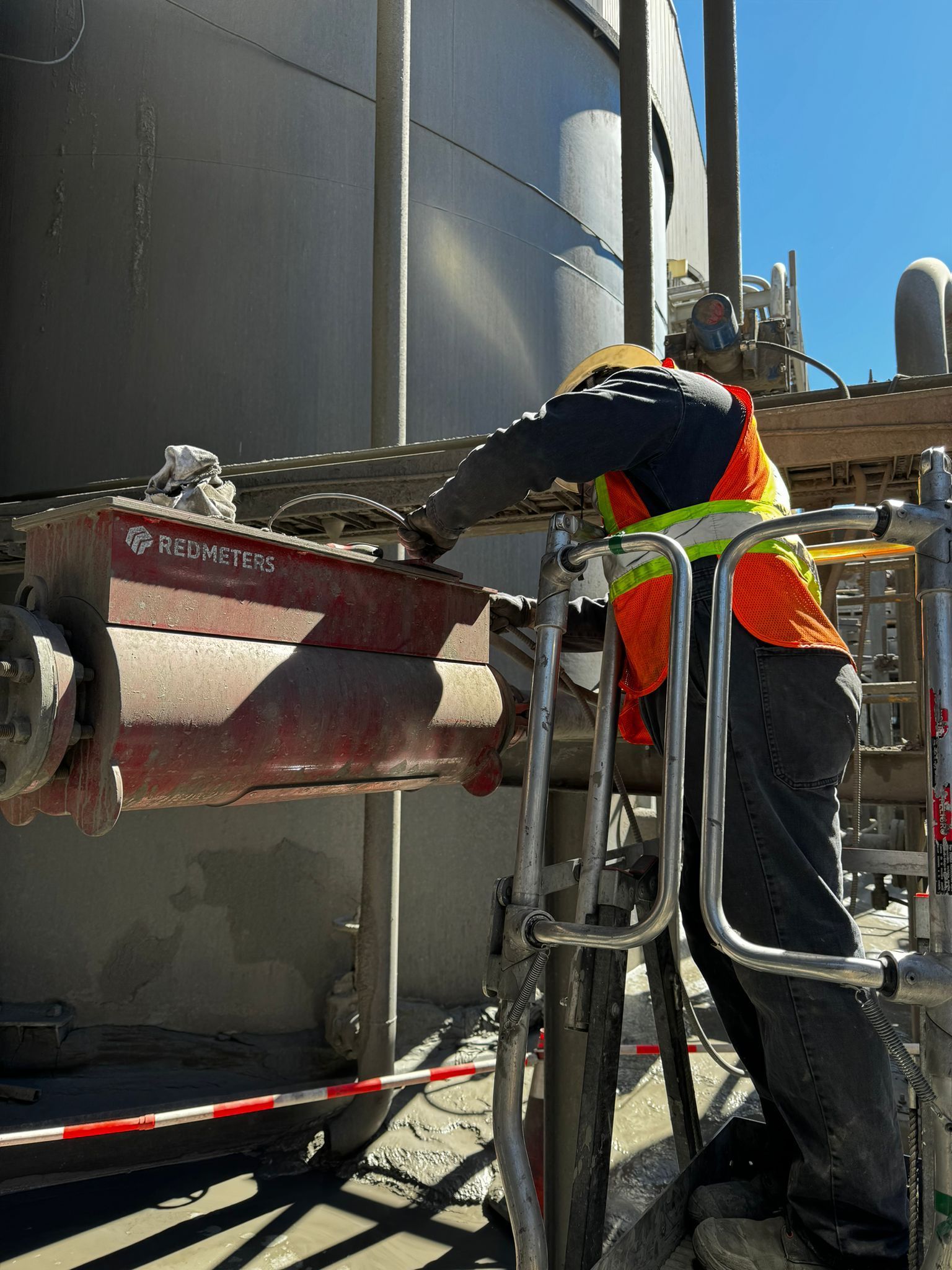 Red Meters Process Control System. A worker in safety gear uses a tool on a large industrial component. The worker stands on a metal scaffolding next to a large tank.