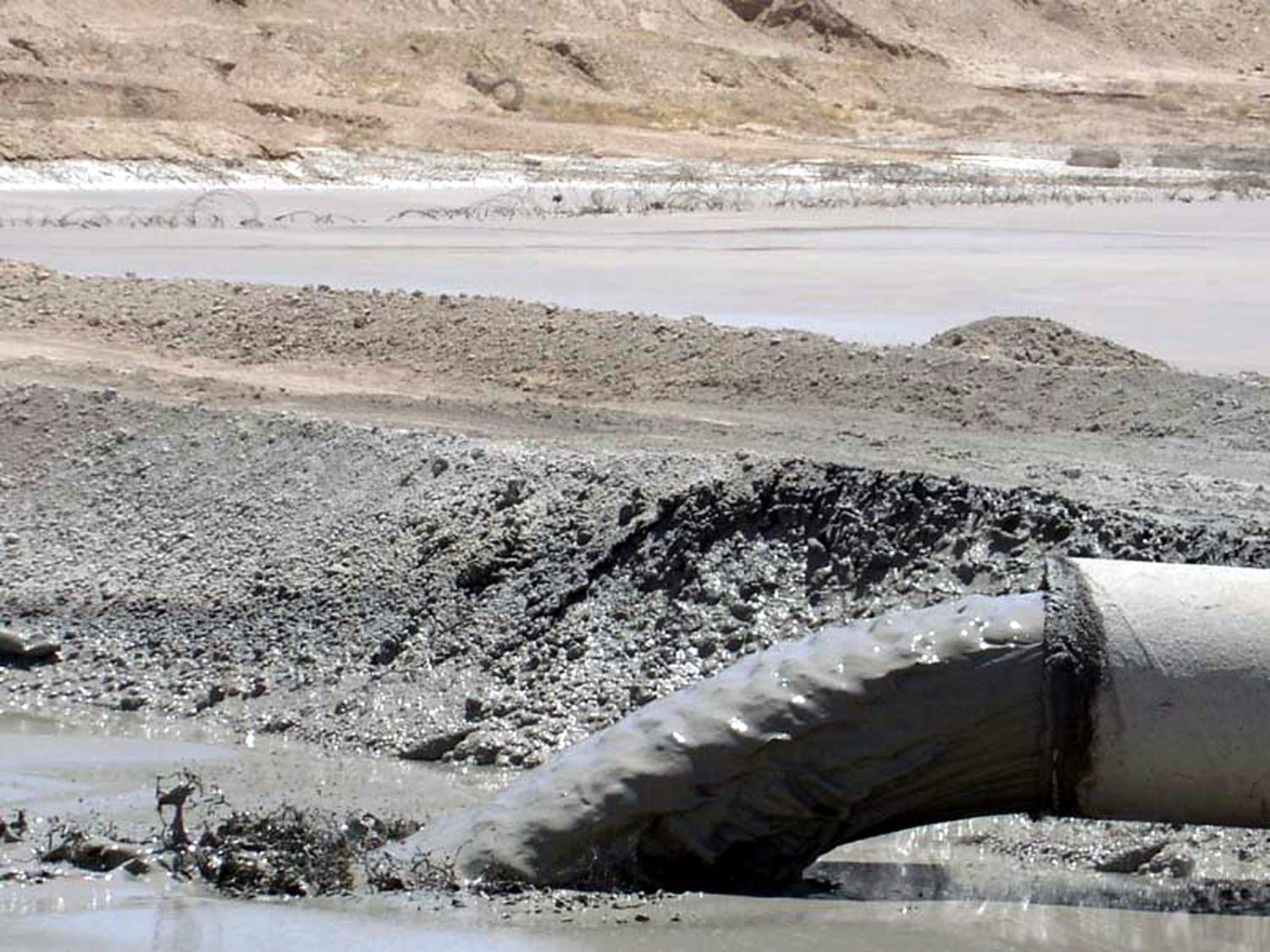 A pipe spews gray sludge into a muddy, desert-like environment with hills in the background.