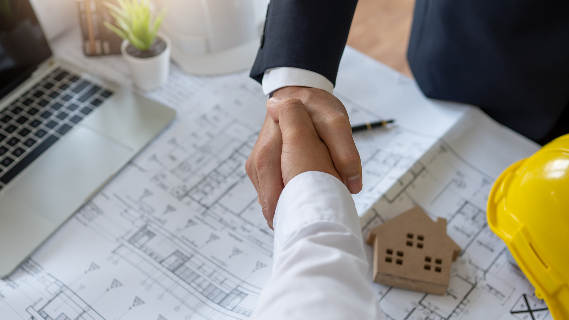 Handshake over architectural blueprints on a desk, with a laptop, plant, and yellow hard hat nearby
