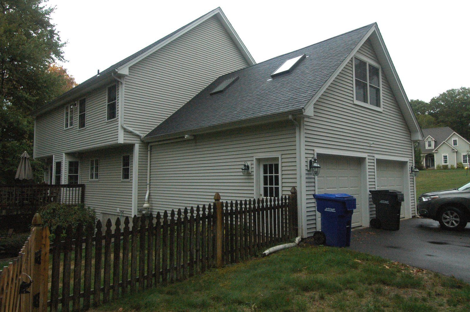 Gray house with a steep roof, white siding, and a fenced yard beside a driveway and trash bins