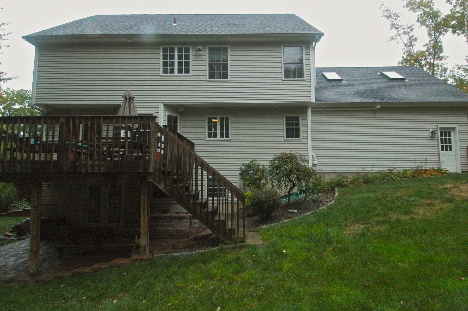 Backyard view of a two-story white house with a wooden deck and stairs on a grassy slope.