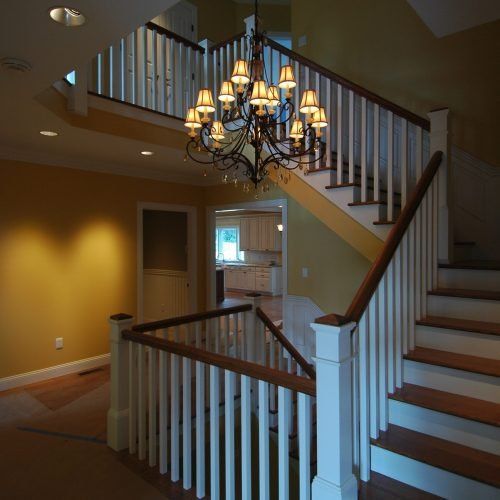 Bright foyer with staircase, chandelier, white railings, and warm yellow walls leading to a kitchen.