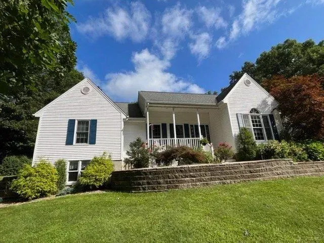White house with gray roof and front porch on a sunny hillside, framed by trees and blue sky