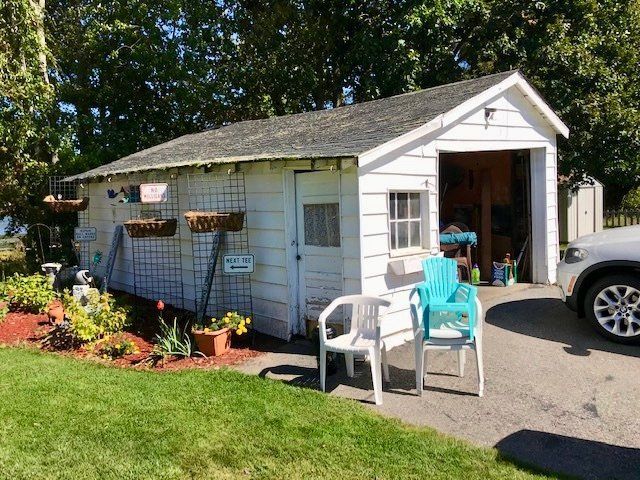 Small white garden shed with chairs and potted plants beside a driveway and lawn