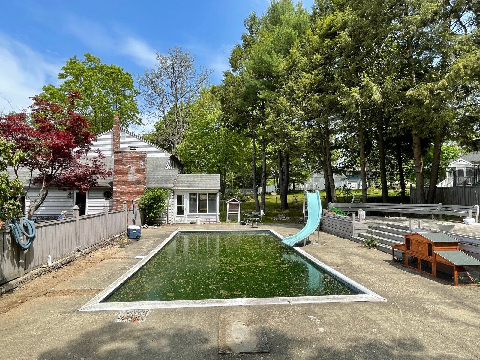 Backyard with a rectangular green pool, slide, patio chairs, and trees under a clear blue sky