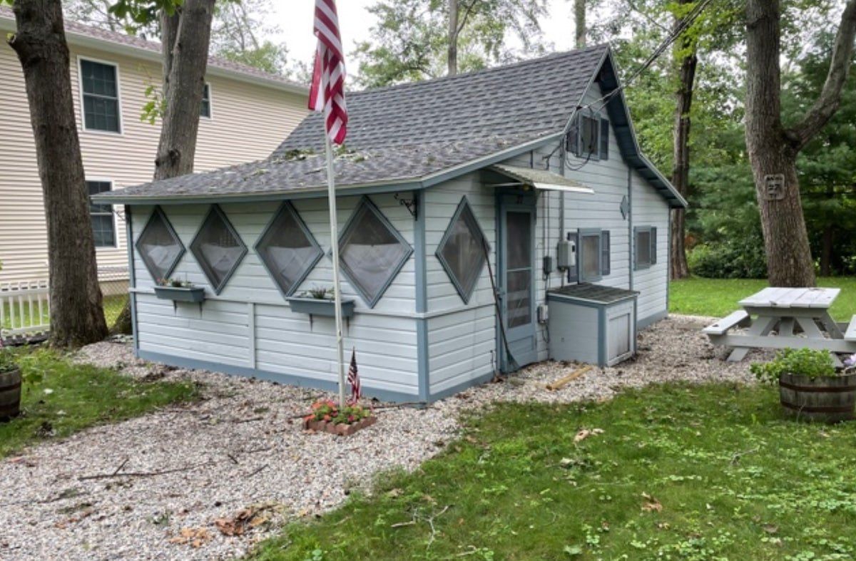 Small light-blue cabin with diamond windows, an American flag, and a picnic table in a wooded yard.