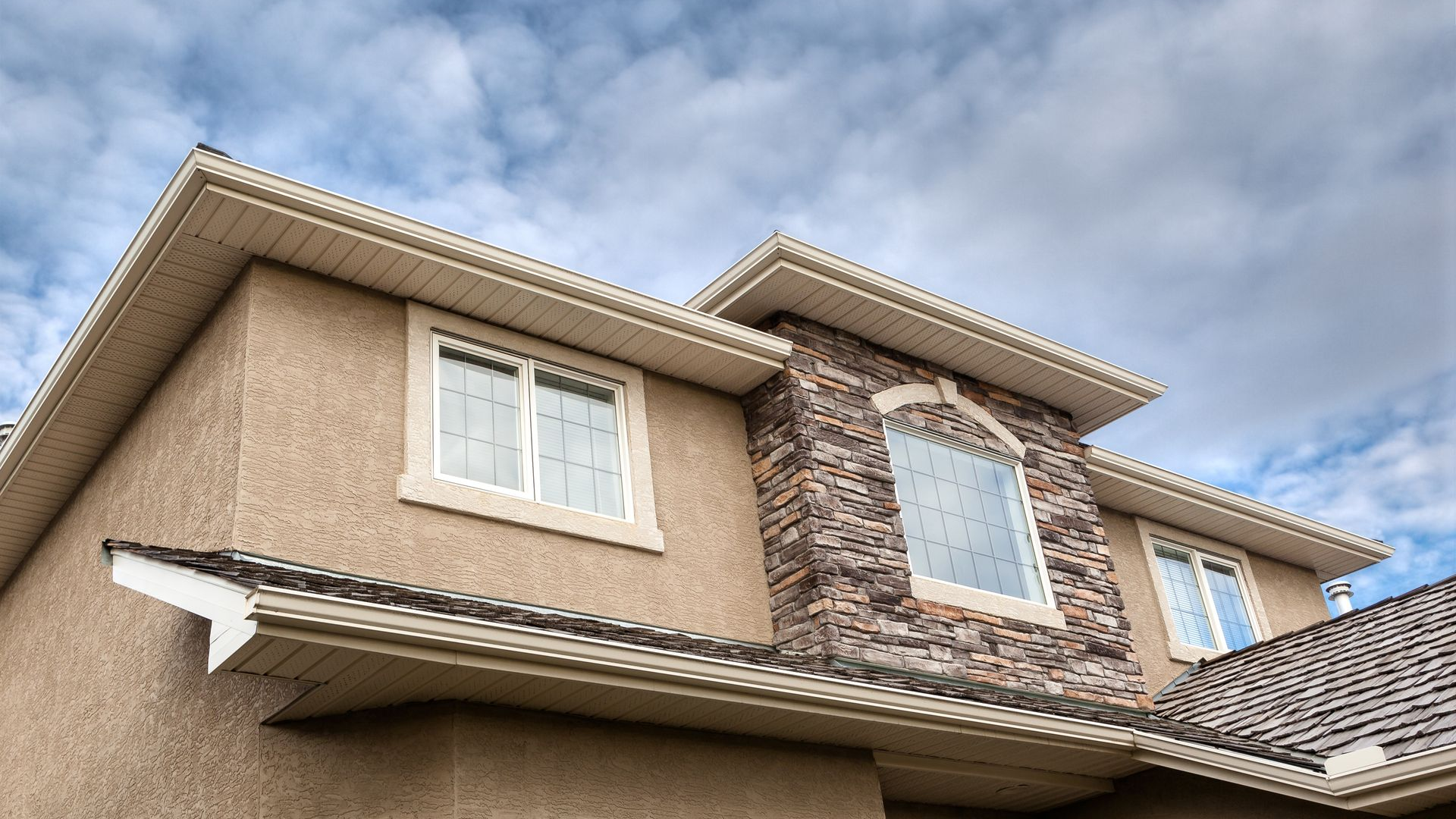 Low-angle view of a beige stucco house with white-trimmed windows and a tiled roof under a cloudy sky