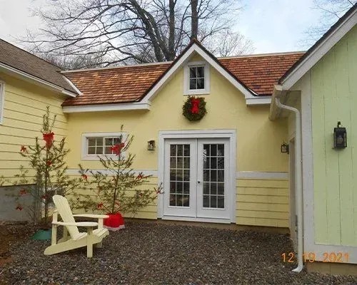 Yellow cottage entry with white French doors, red wreath, and a beige bench on a gravel courtyard.