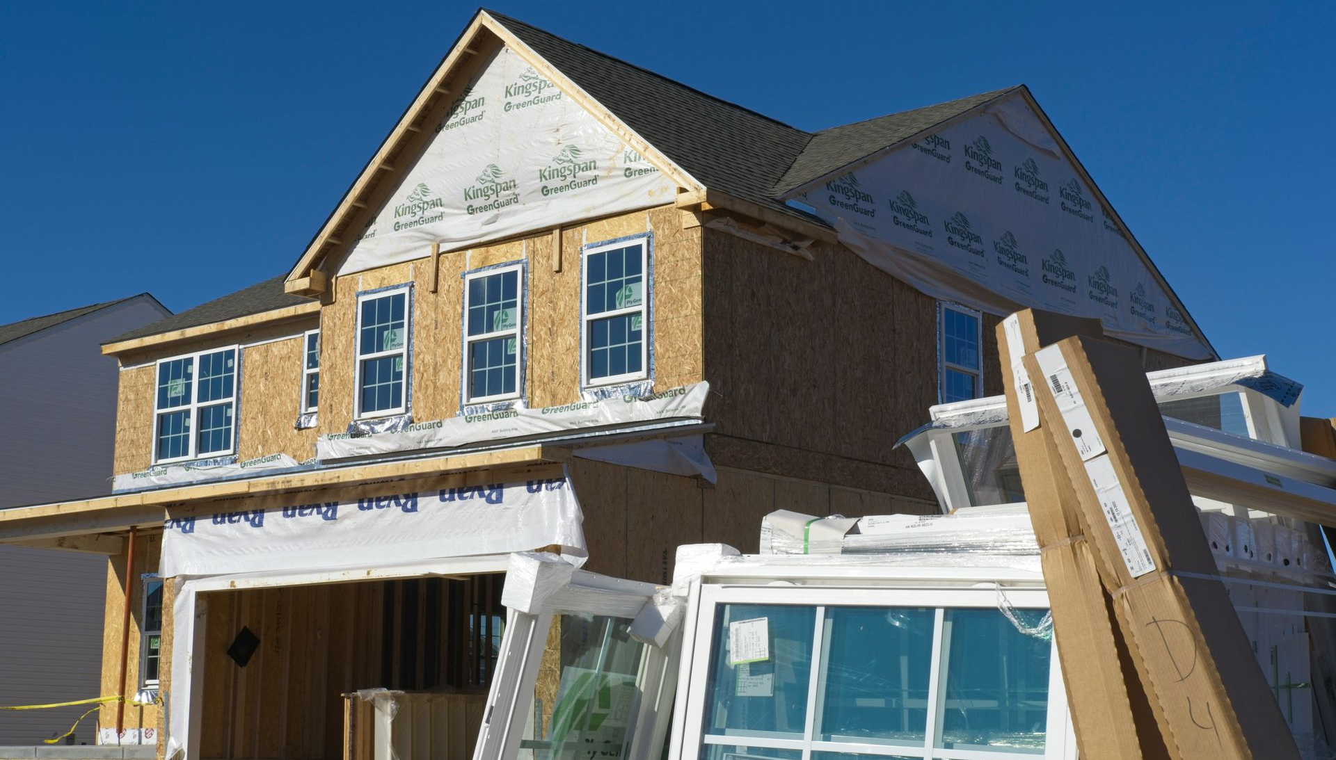 House under construction with exposed wood framing, white siding, and a steep roof under a clear blue sky