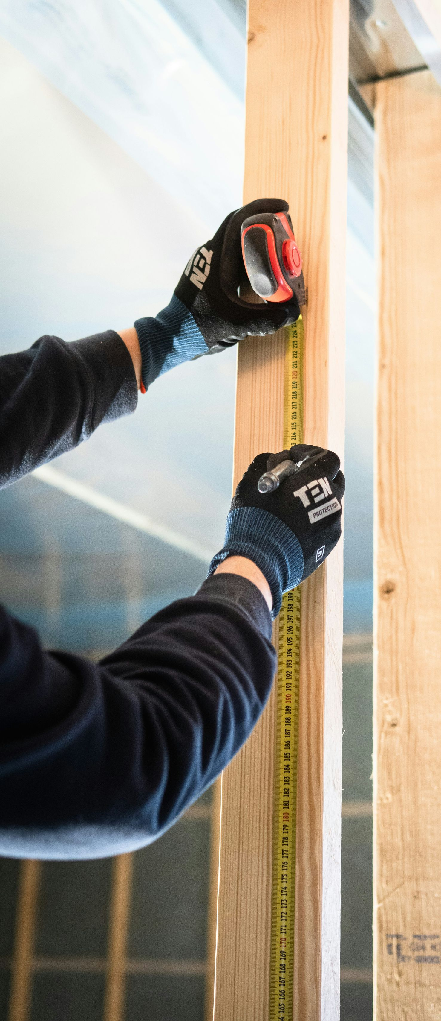 Gloved hands measure a wooden stud with a tape measure and pencil in a construction setting