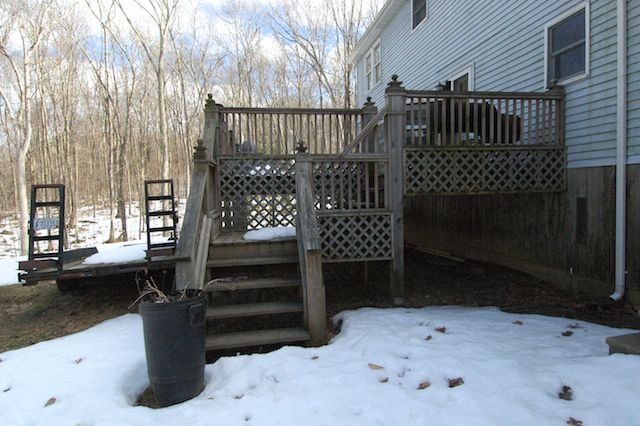 Snowy backyard deck with wooden stairs, railing, and a black trash bin nearby