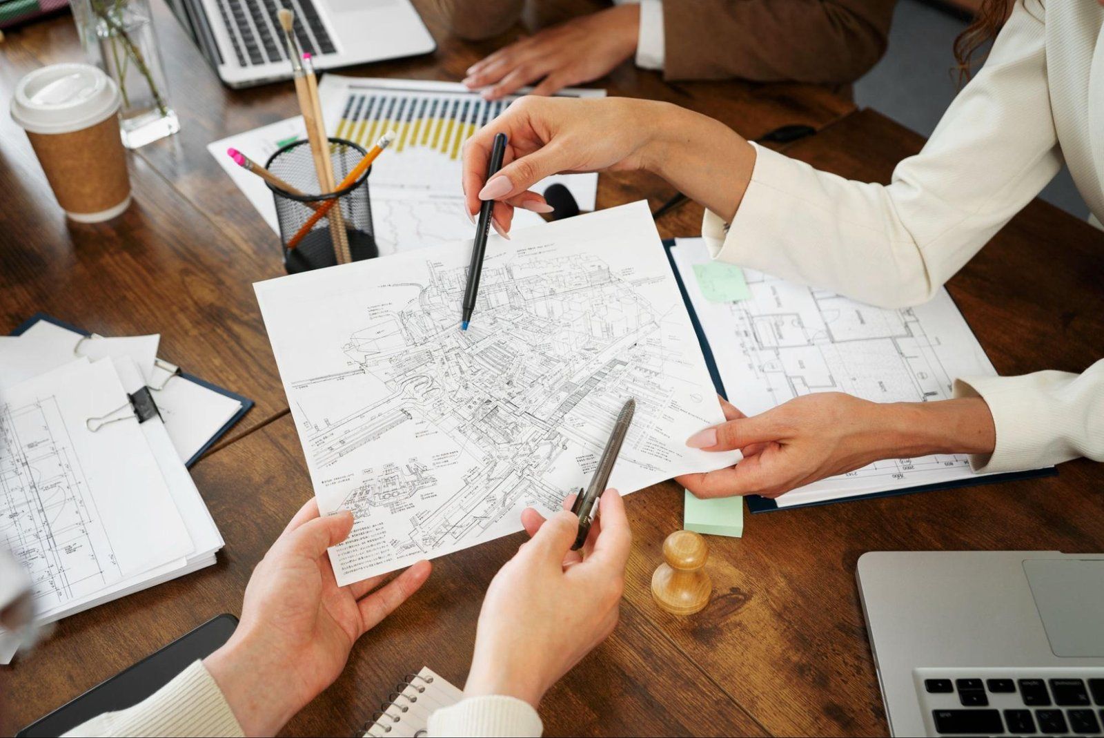 Hands pointing at a sketch-covered table with papers, pencils, coffee, and a laptop in a meeting setting