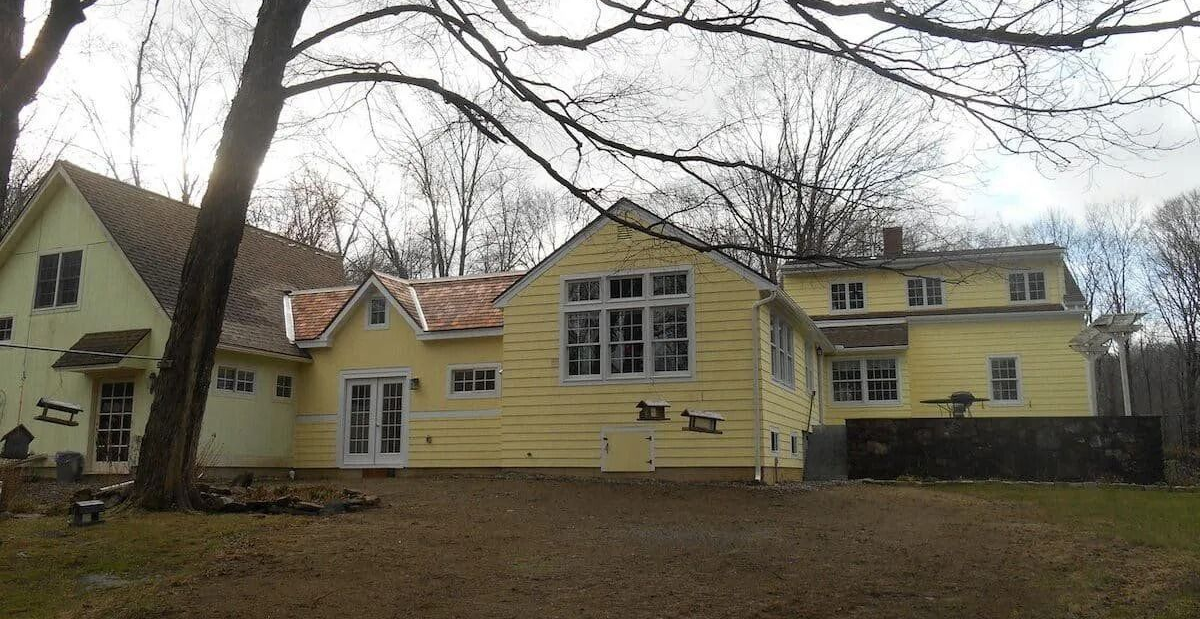 Yellow house with large windows and leafless trees in a yard under an overcast sky