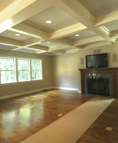 Empty living room with hardwood floors, coffered ceiling, windows, fireplace, and TV mounted above the mantel