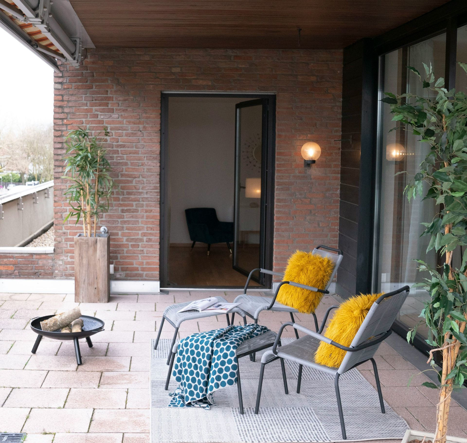 Cozy brick patio with woven chairs, yellow cushions, a small fire pit, and an open doorway to a dim room