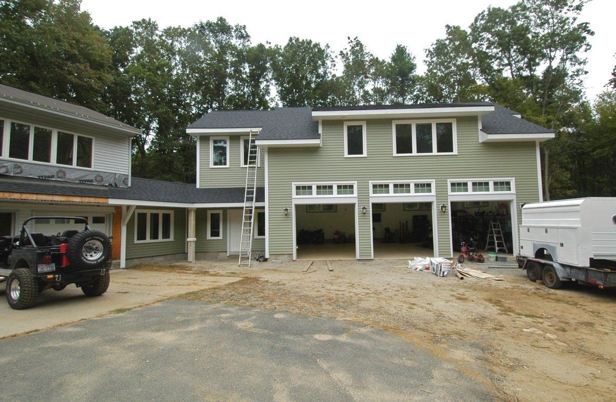 House under construction with three garage openings, ladders, and work vehicles in the driveway.
