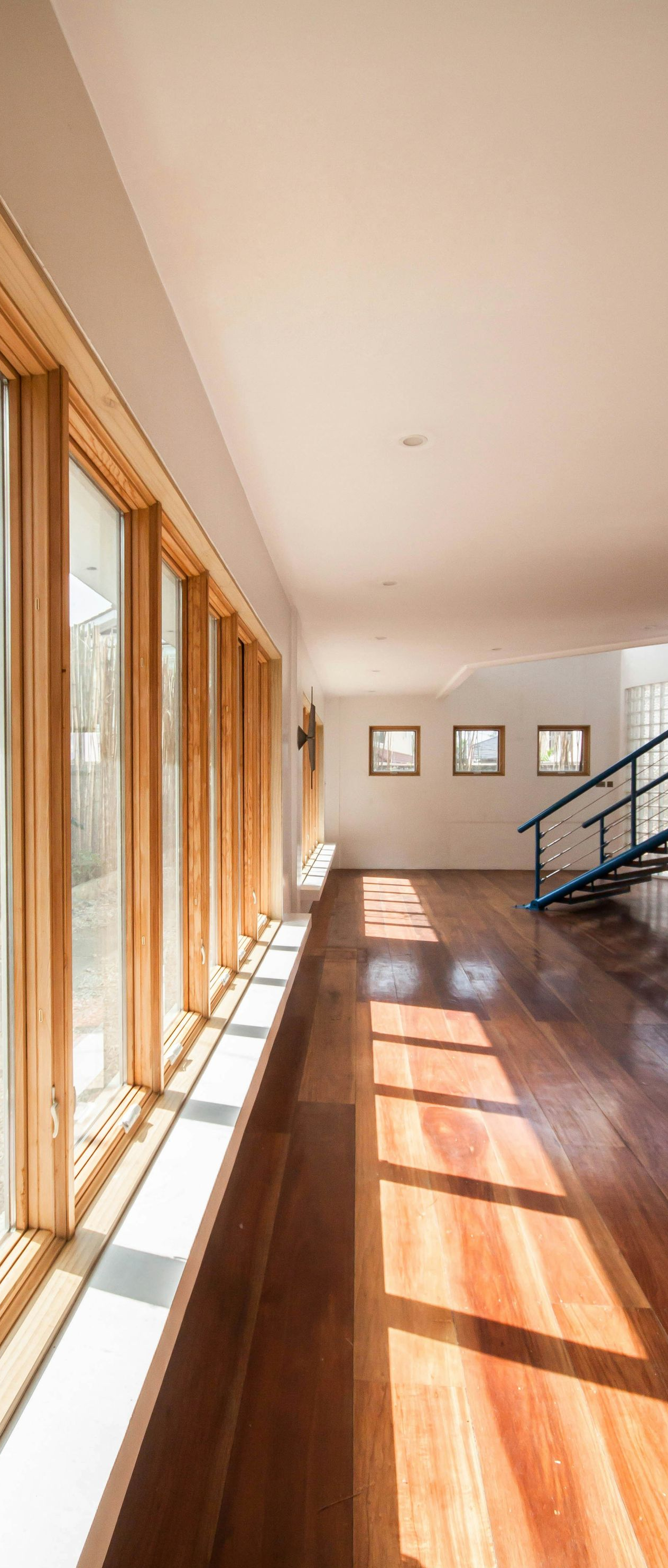 Sunlit wooden hallway with large windows, glossy floor, and black staircase railing