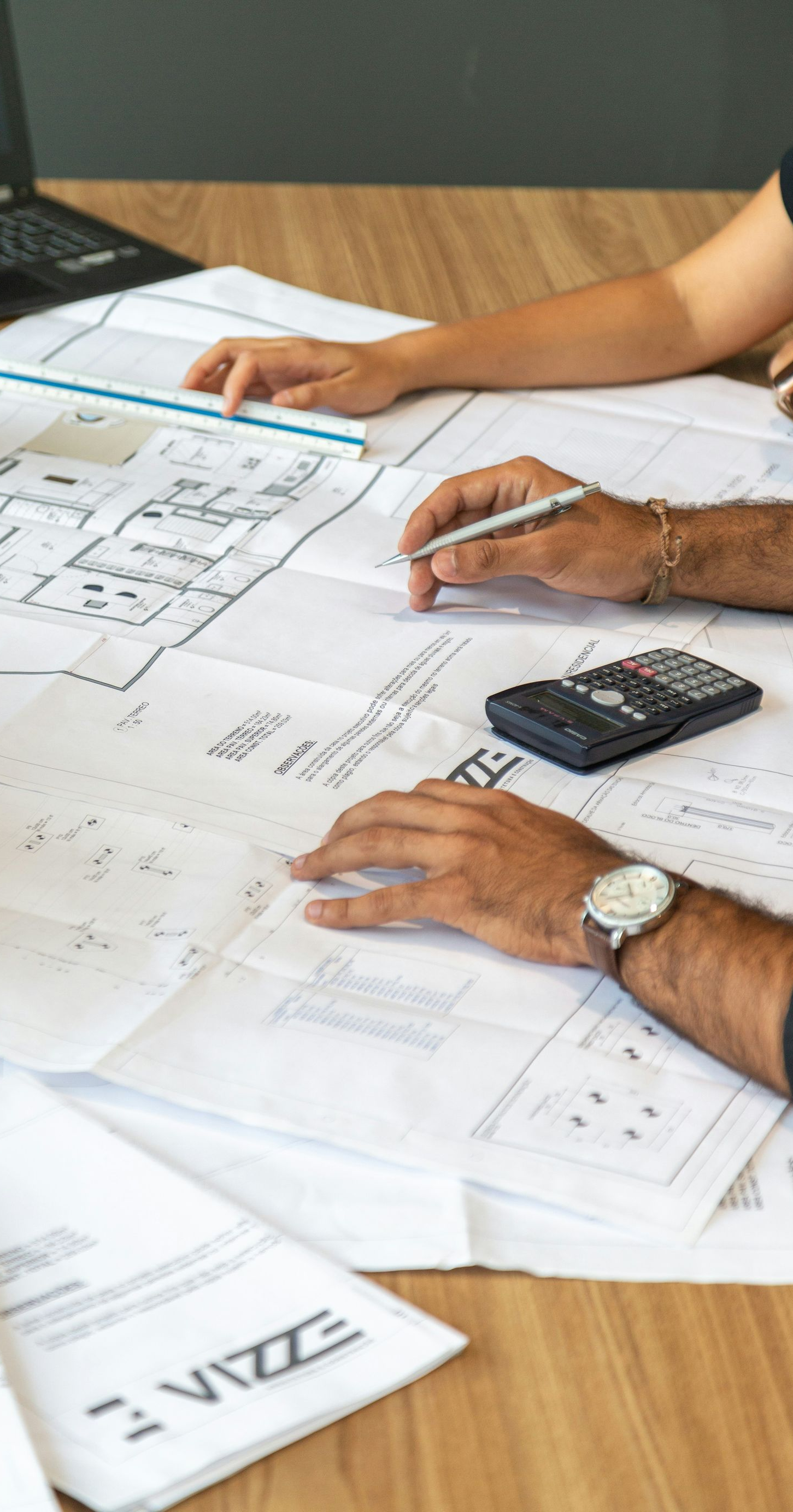 Hands reviewing architectural plans on a wooden table with a calculator and ruler