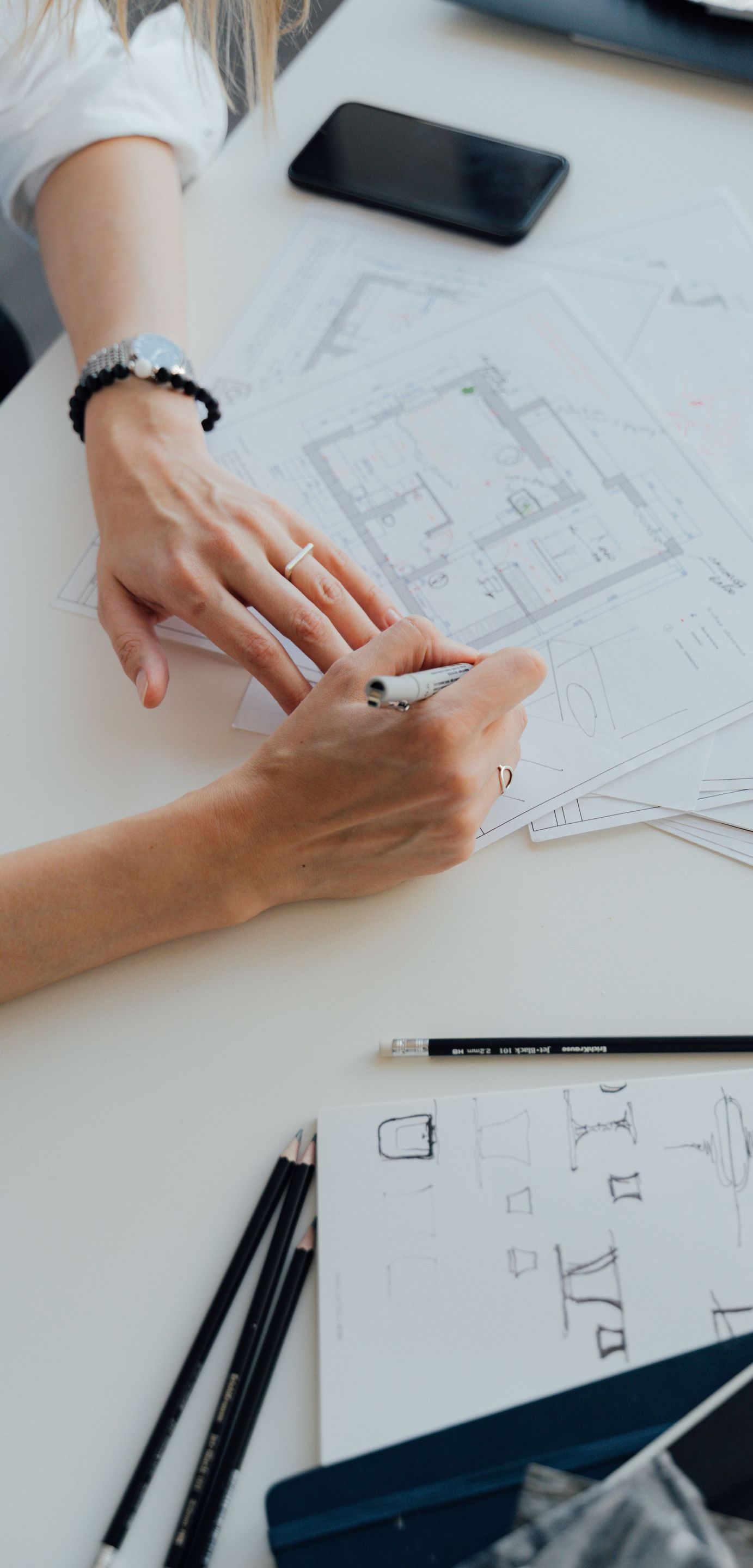 Hands sketching a design on paper beside a smartphone and drafting tools on a desk