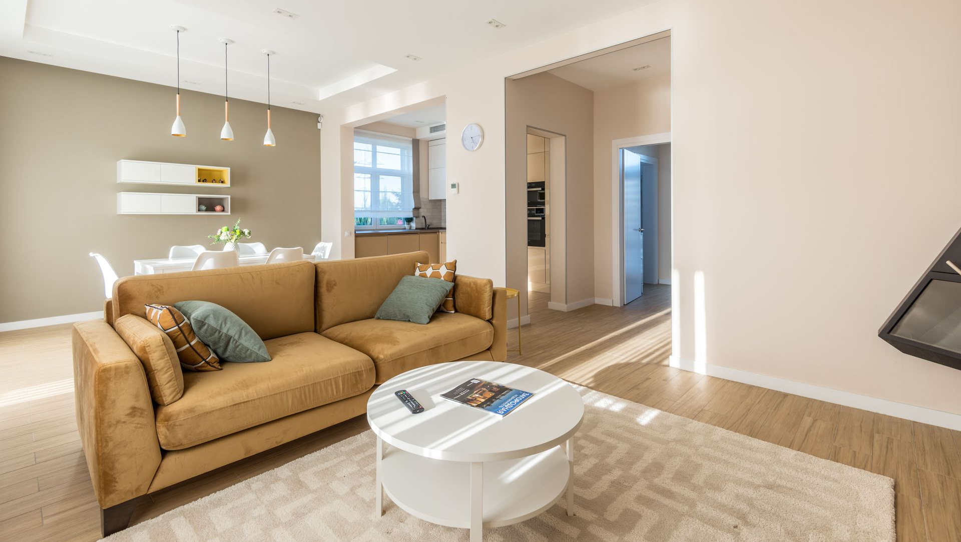 Bright living room with tan sofa, white coffee table, rug, and open kitchen in the background